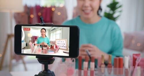 Woman sitting at a table in the background, with a phone on a tripod recording video in the foreground.